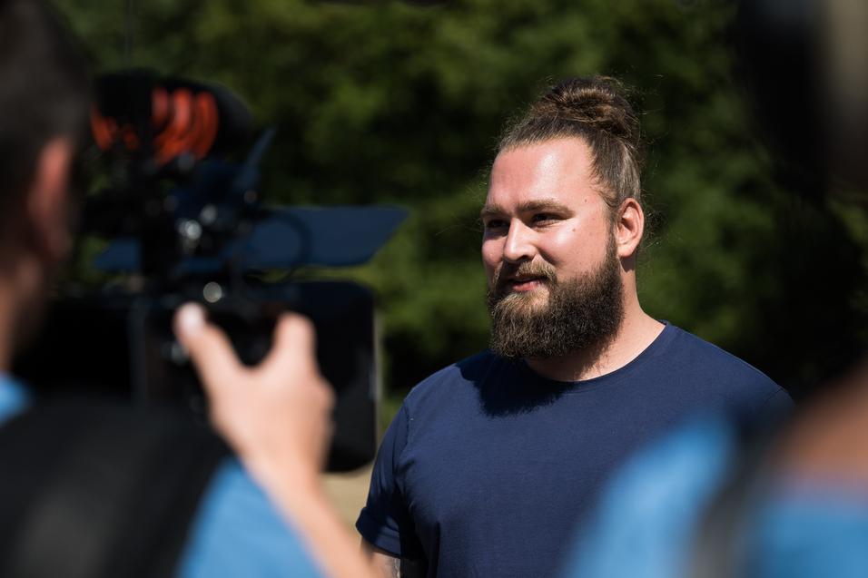 Mann mit Bart und Dutt steht vor einer Kamera und spricht, während ein weiteres Kamerateam im Hintergrund sichtbar ist.